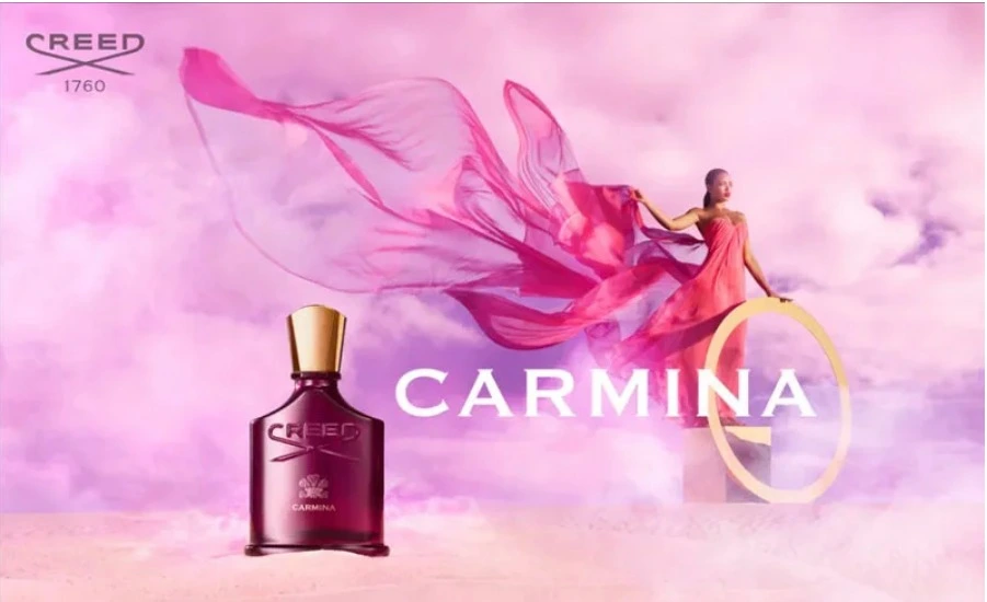 Anúncio do perfume Carmina da Creed com frasco vermelho e tampa dourada em destaque, ao lado de uma mulher com vestido rosa esvoaçante em cenário etéreo com tons rosados, transmitindo luxo e feminilidade.