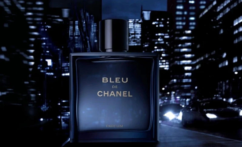 Frasco do perfume Bleu de Chanel Parfum em destaque, com design azul escuro elegante, posicionado diante de um cenário urbano noturno com prédios iluminados e carros, transmitindo sofisticação, modernidade e masculinidade.