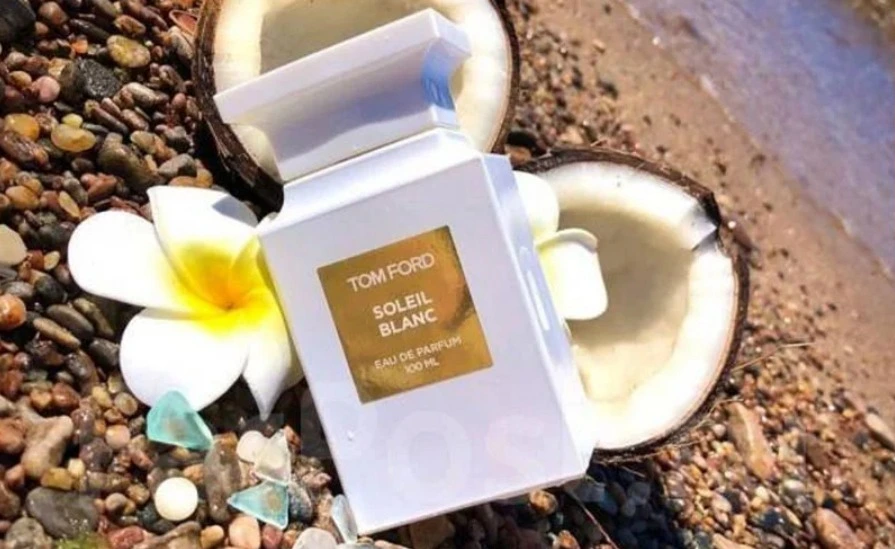 Perfume Soleil Blanc Tom Ford em frasco branco apoiado sobre pedras na praia, com coco aberto ao fundo e flor branca ao lado, evocando um cenário tropical sofisticado e ensolarado.