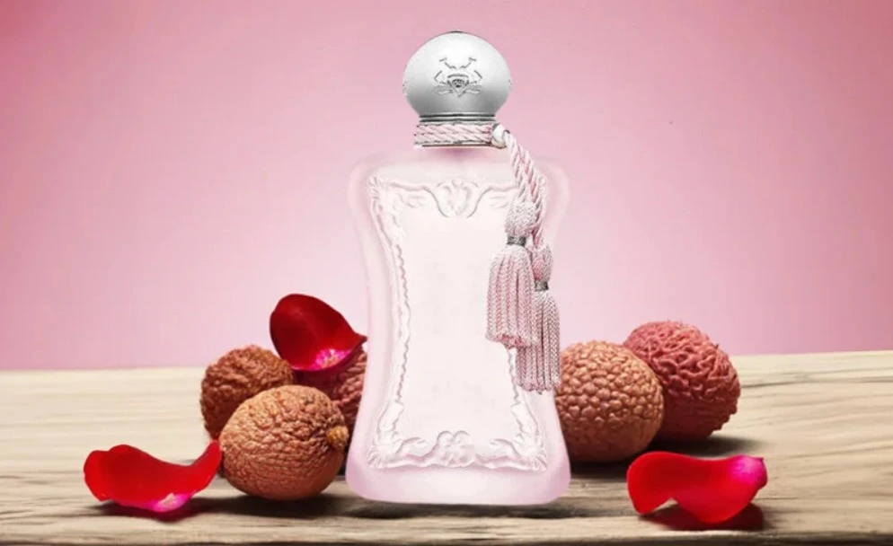 Frasco do perfume Delina Parfums de Marly em tom rosado com tampa prateada e detalhe de tassel, posicionado sobre superfície de madeira, cercado por lichias e pétalas de rosa, em fundo rosa suave que transmite delicadeza, feminilidade e sofisticação.