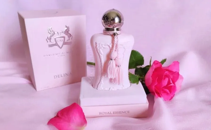 Frasco do perfume Delina Parfums de Marly em tom rosado com tampa prateada e detalhe de tassel, acompanhado da caixa ao fundo e rosas ao redor, em cenário suave que transmite feminilidade, elegância e romantismo.