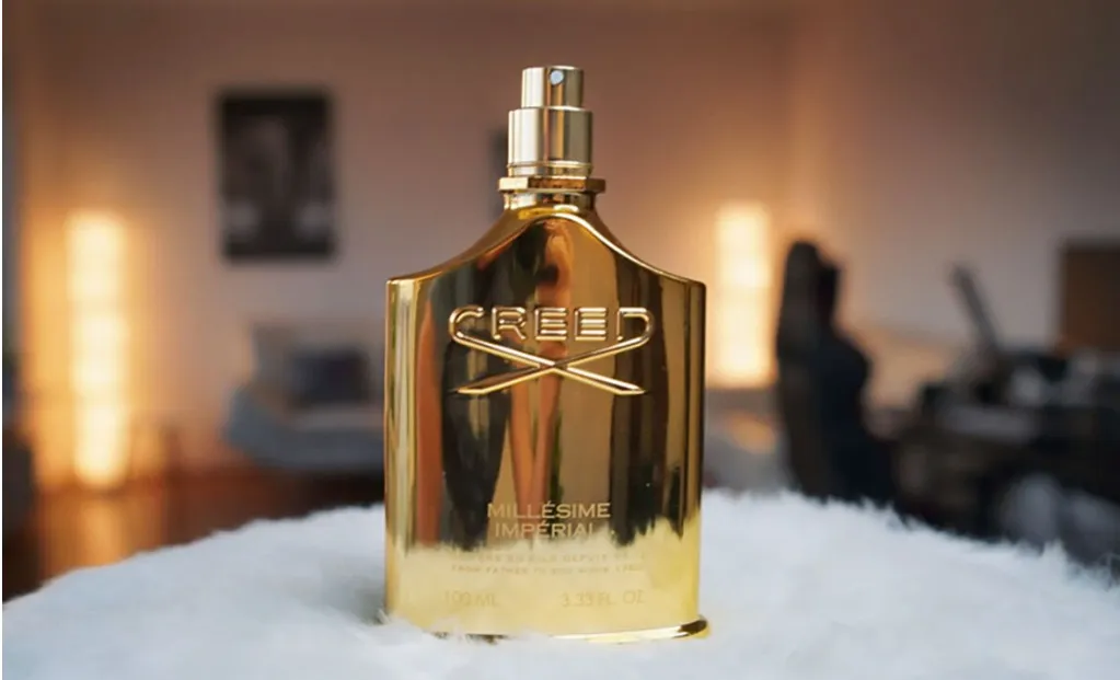 Frasco do perfume Millésime Impérial Creed em acabamento dourado metálico, posicionado sobre superfície macia com fundo desfocado de ambiente sofisticado, transmitindo luxo e elegância.