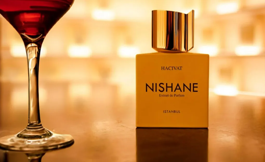Frasco do perfume Hacivat Nishane em cor dourada com tampa metálica brilhante, posicionado sobre mesa ao lado de uma taça de drink vermelho, com fundo iluminado que transmite luxo, sofisticação e elegância noturna.