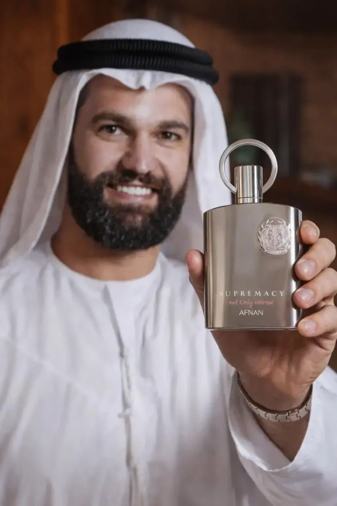 João da Barba sorrindo vestido com roupa árabe branca segurando o perfume Afnan Supremacy Not Only Intense próximo à câmera, destacando fragrância masculina intensa, sofisticada e marcante dos perfumes árabes